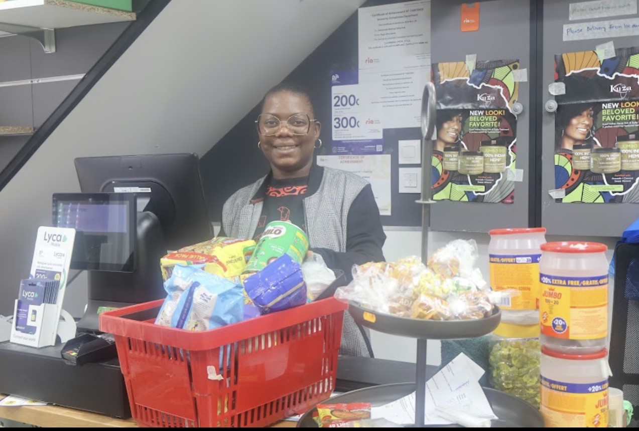 Smiling TEGA AFRO STORE shopkeeper at the till with a basket of African groceries